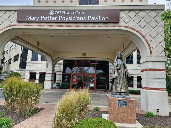 Bronze statue of a nun holding a crucifix in front of a building entrance with a sign overhead that reads “OSF HealthCare Mary Potter Physicians Pavilion.”