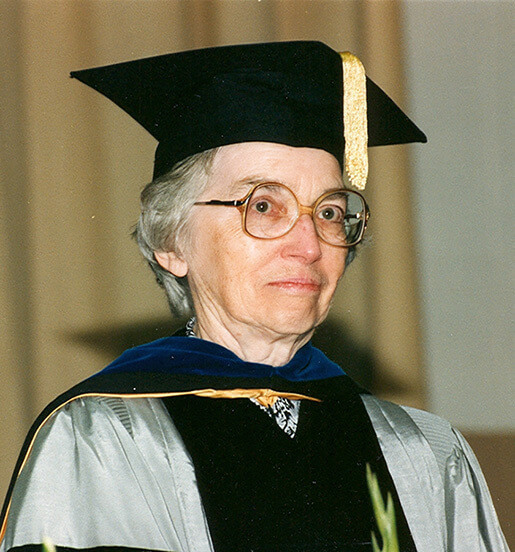 A woman with glasses wears academic regalia, including a black mortarboard with a gold tassel and a silver gown with a dark blue and gold stole.