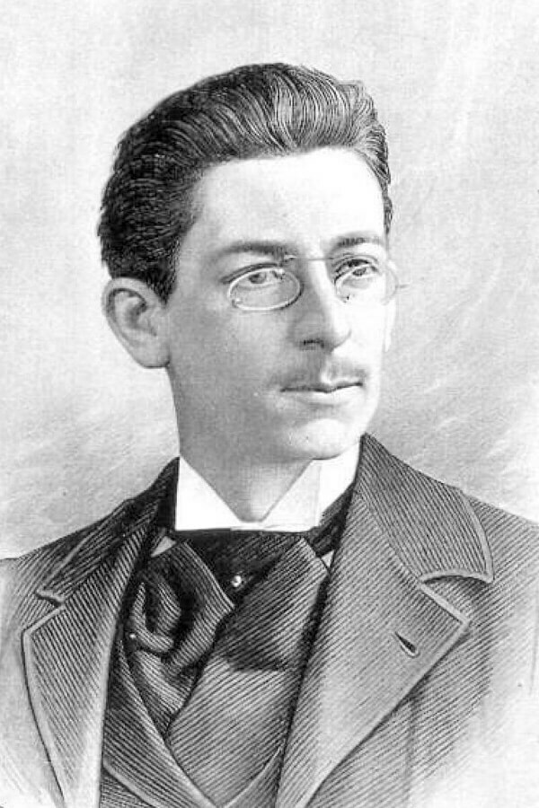 Black and white portrait of a man with neatly styled dark hair, a mustache, and round spectacles. He wears a formal suit, vest, white shirt, and a dark cravat, gazing off-center with a neutral expression.