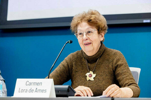Carmen Lora, in glasses & brown sweater, smiles while speaking into a microphone, wearing a star necklace.