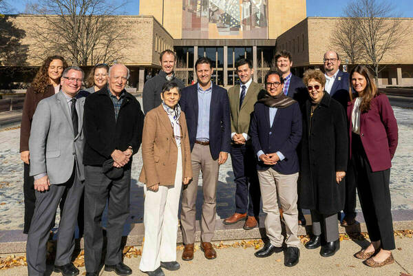 Eight men and five women smile in various business attire in front of Notre Dame's Hesburgh Library on a sunny day.