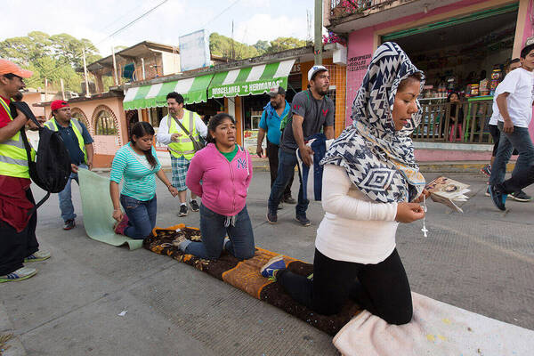 Three women kneel on blankets in a street procession. Surrounding them are several men walking and moving the blankets as they make progress.