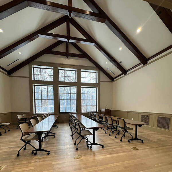 Meeting room with vaulted ceiling, dark wood beams, light walls, long tables, and a podium in the corner. Large windows show bare trees.