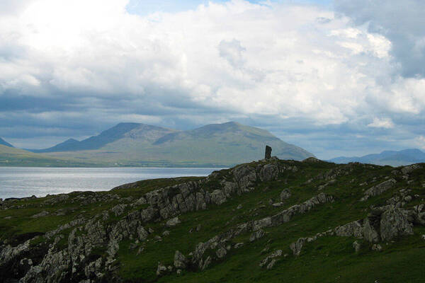Rocky, grassy hills with a tall dark standing stone overlooking a calm lake and distant green mountains under a cloudy sky.