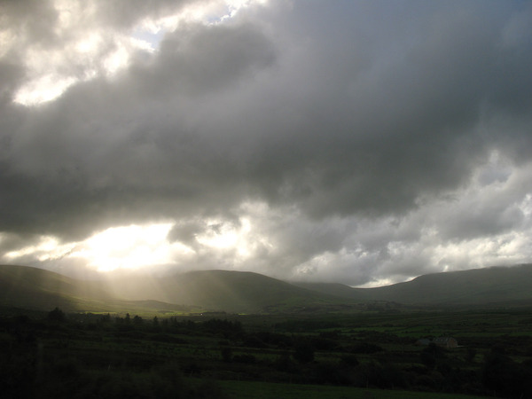 Dark, stormy clouds fill the sky above rolling green hills, with bright sunlight breaking through and illuminating the valley.