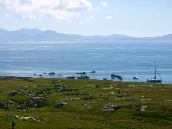 Blue sea with many boats near a rocky, green shore and distant mountains under a partly cloudy sky.