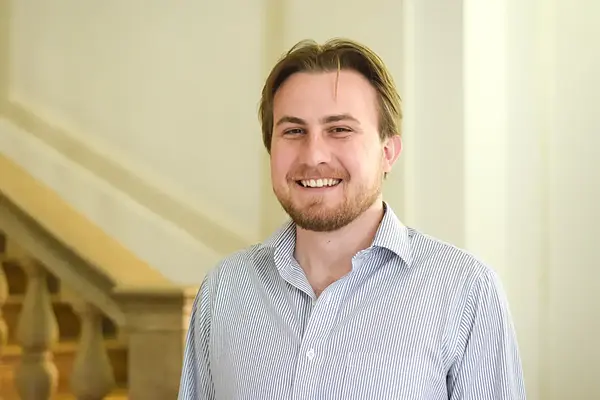 Young man with light brown hair and beard, wearing a blue and white striped shirt, smiles broadly in a hallway.