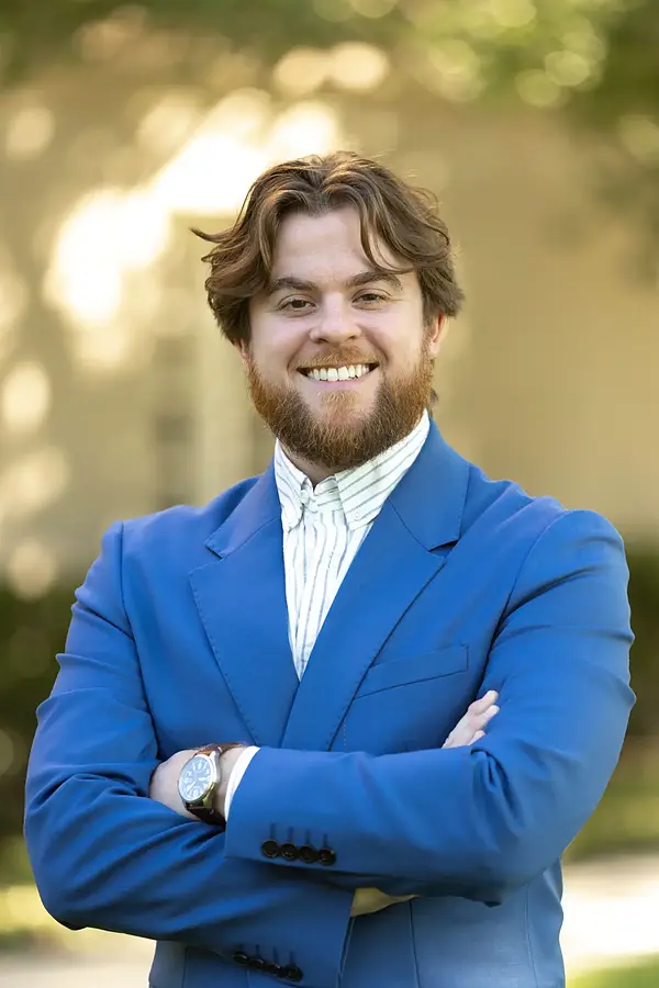 Man with brown hair and beard, wearing a blue suit and striped shirt, smiling broadly with arms crossed outdoors.